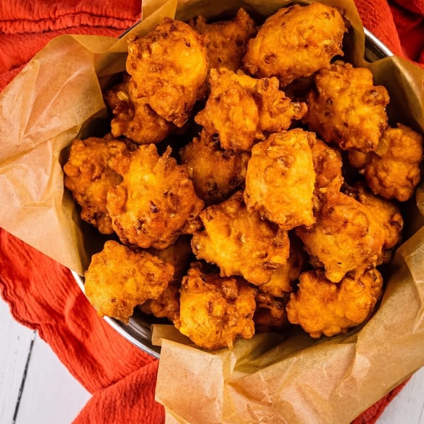 Golden-brown fried fritters, possibly hush puppies or corn fritters, in a metal bowl lined with parchment paper.